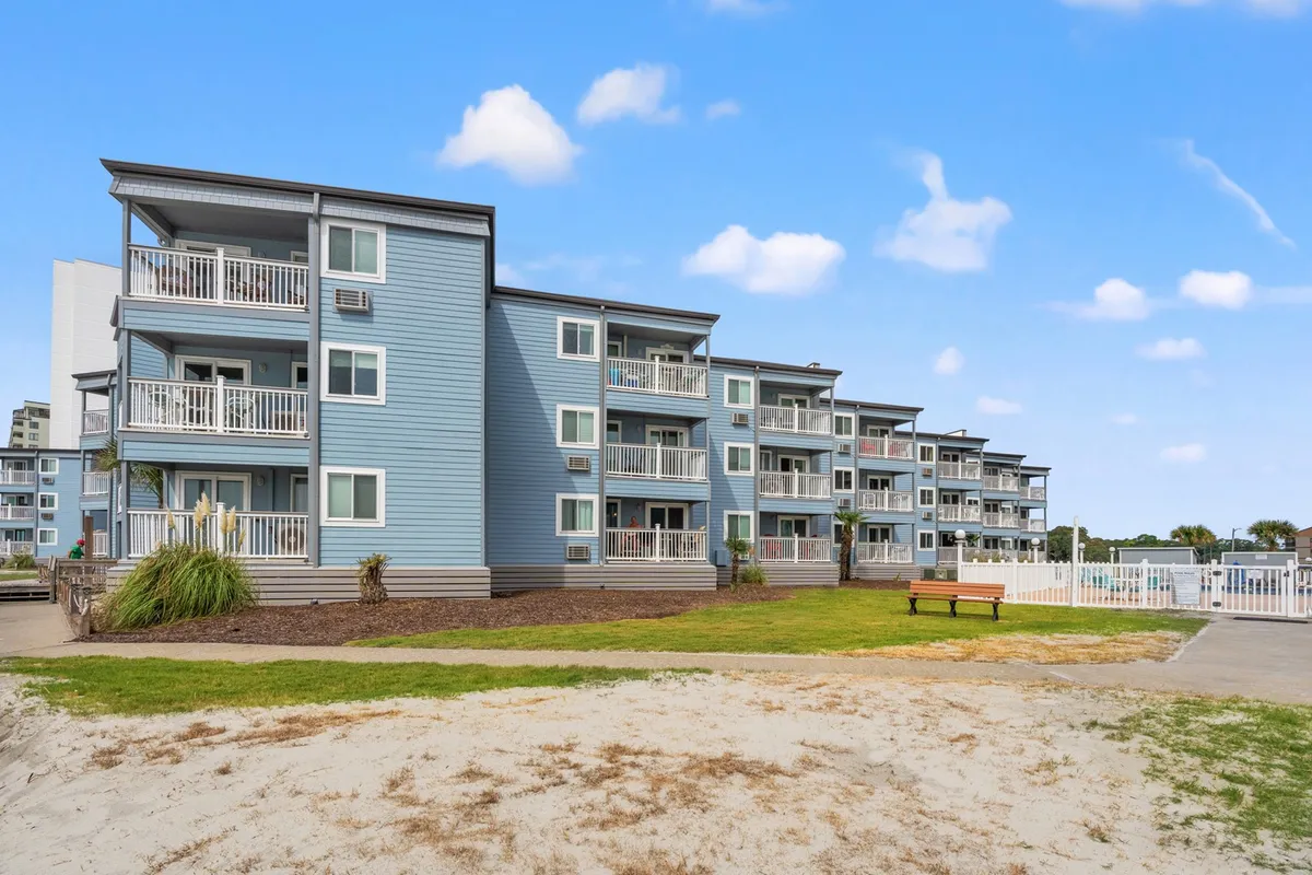 This image shows the exterior of blue apartment buildings surrounded by a green lawn, a bench, and fencing. The scene exudes a welcoming and peaceful atmosphere, with hints of outdoor relaxation and community. The open outdoor spaces suggest a safe, inviting environment perfect for extended stays during the quieter winter months.
