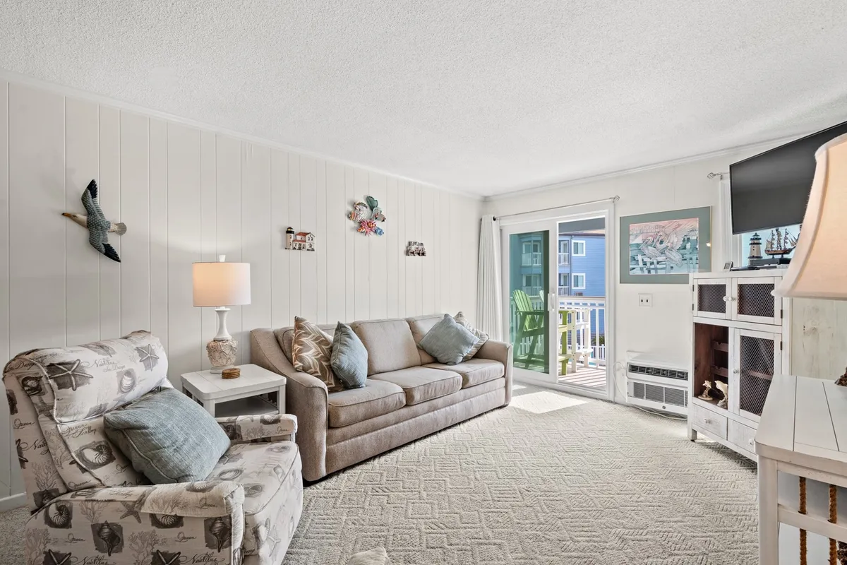 A comfortable living room featuring a plush sofa and armchair, a stylish lamp, and a sliding glass door leading to a balcony. The room is bathed in natural light, offering a glimpse of the outdoors and suggesting access to fresh breezes and views.