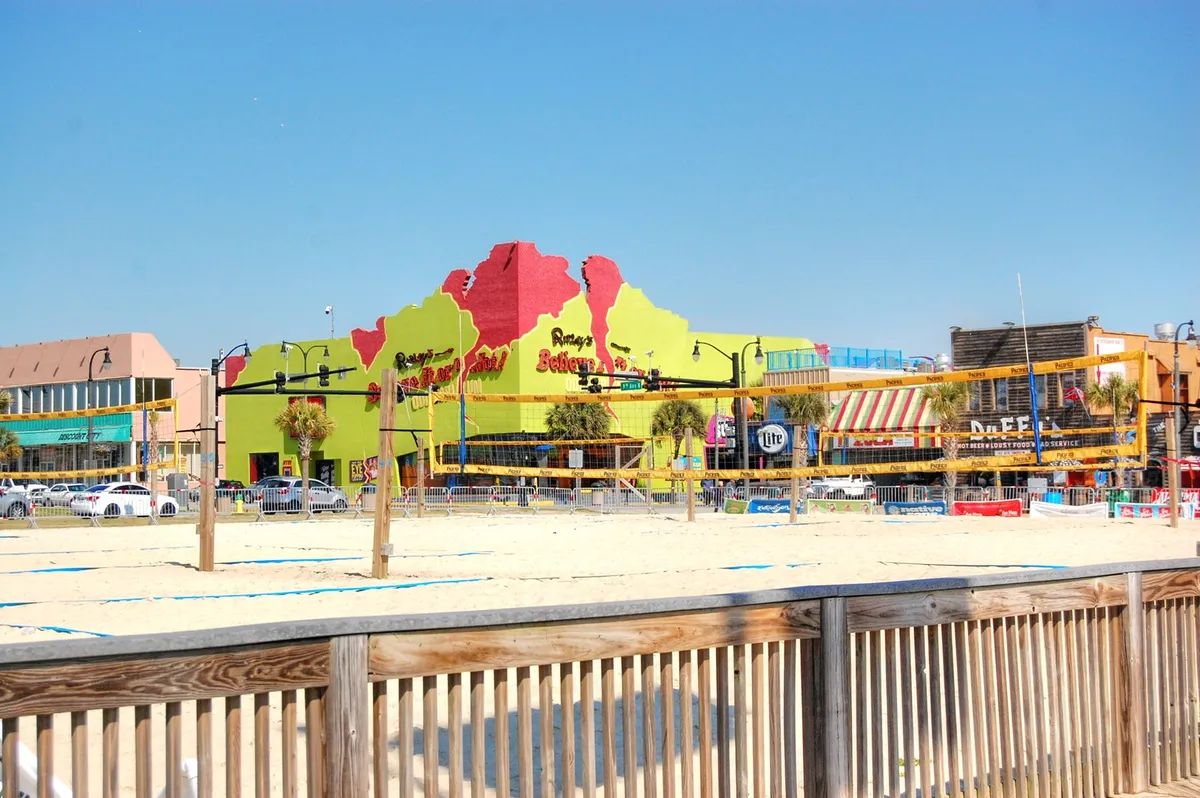 The image displays a vibrant exterior with a colorful building facade, beach volleyball courts, palm trees, and outdoor seating areas. The lively scene suggests energetic activities, cultural experiences, and social opportunities available nearby.