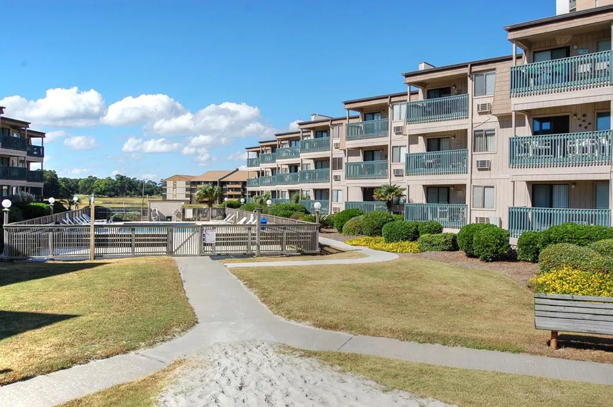 A welcoming exterior view with landscaped gardens, balconies overlooking the scene, and a pathway leading toward the pool area. The environment encourages group strolls, poolside relaxation, and community events in a tranquil yet inviting setting.