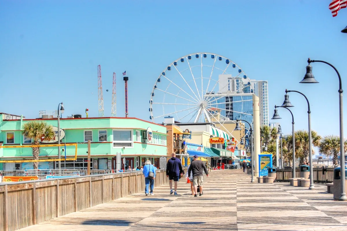 A colorful beachfront boardwalk featuring a towering Ferris wheel, vibrant storefronts, and palm trees. The lively scene hints at the nearby SkyWheel, local shops, and dining options—representative of the area’s energetic spirit and variety of attractions.