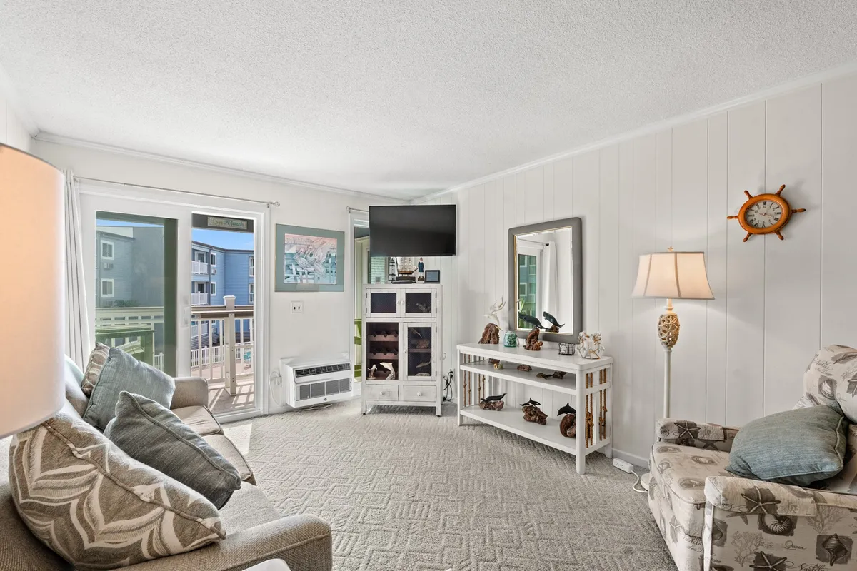 A sunlit living room featuring a comfortable sofa with patterned cushions, a sleek console table decorated with tasteful items, a flat-screen television, and expansive windows letting in abundant natural light. The space exudes a sense of welcome, comfort, and relaxation.
