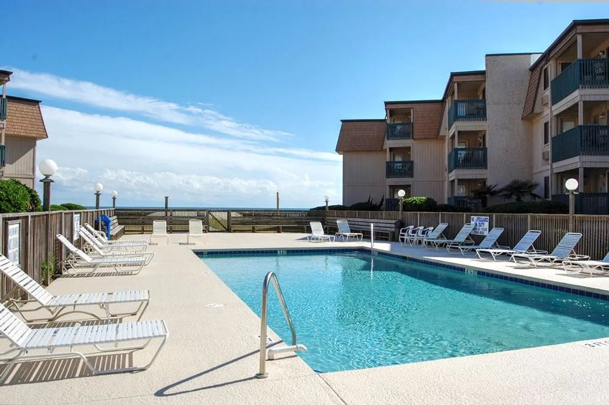 A vibrant outdoor swimming pool surrounded by lounge chairs and nearby buildings, with glimpses of the ocean in the background. People are enjoying swimming, sunbathing, and relaxing poolside, creating a laid-back and leisurely atmosphere.