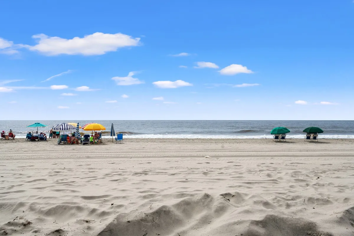 A picturesque beach scene with umbrellas, seaside chairs, ocean waves, and sand dunes. People are sunbathing, swimming, and playing games, soaking up the carefree beach atmosphere with sunset views in the distance.
