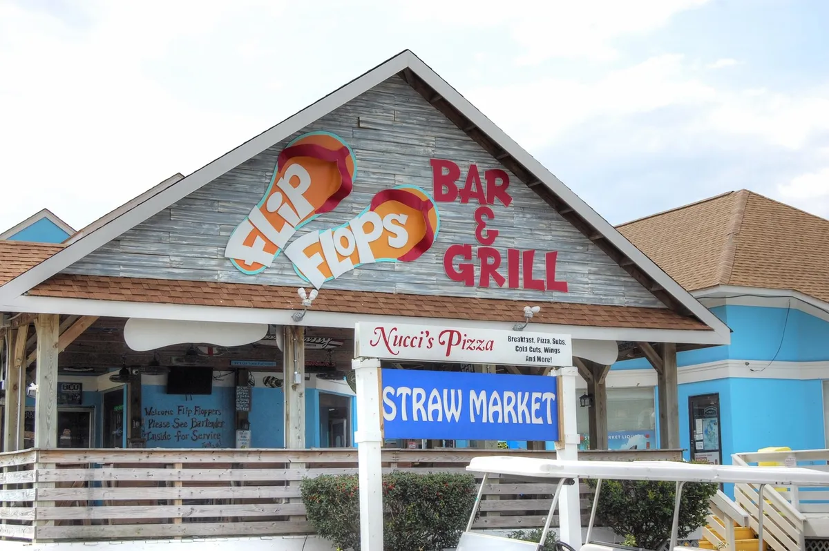 The entrance to a colorful plaza featuring signs for Flip Flops Bar & Grill, Nucci's Pizza, and the Straw Market, with outdoor seating and a casual, festive street scene. The setting suggests a hotspot for dining, socializing, and enjoying local events with friends.