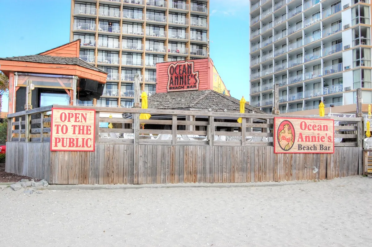 This image features the iconic Ocean Annie's Beach Bar, complete with its recognizable signage and inviting seating area shaded by umbrellas. The lively beachfront atmosphere is visible, suggesting a hub for social gatherings, live music, and ocean views—capturing the essence of local nightlife and relaxation.